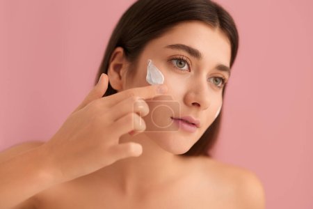 Photo for Young female with perfect smooth skin applying moisturizing cream on face looking away against pink background - Royalty Free Image
