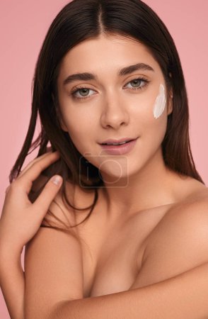 Photo for Self assured young topless female model with long dark hair and facial cream on cheek, covering breast with arms and looking at camera against pink background - Royalty Free Image
