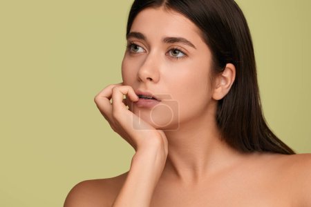 Photo for Pensive young woman with bare shoulders and dark hair touching lips and looking away while thinking about skin care routine against green background - Royalty Free Image