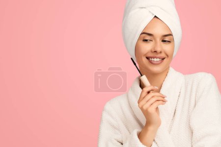 Téléchargez les photos : Joyeux jeune femme en peignoir blanc et serviette sur la tête tenant la brosse de mascara lors de l'application de maquillage sur fond rose - en image libre de droit