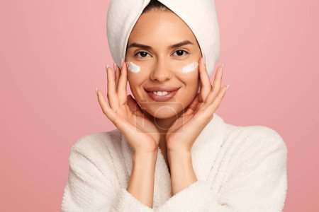 Photo for Smiling young female model in soft bathrobe and towel on head applying moisturizing cream on cheeks while standing against pink background - Royalty Free Image