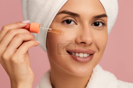 Photo for Crop young female with perfect skin and towel on head smiling and looking at camera while applying moisturizing serum oil on face after bath - Royalty Free Image