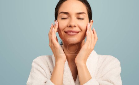 Téléchargez les photos : Jeune femme souriante détendue en peignoir appliquant un produit cosmétique sur le visage avec les mains pendant la procédure de soins de la peau après spa sur fond turquoise - en image libre de droit