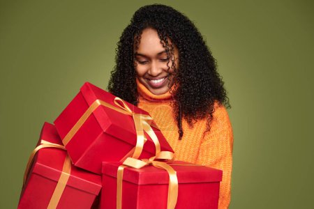 Téléchargez les photos : Femme afro-américaine joyeuse en pull chaud avec des cheveux bouclés portant des boîtes-cadeaux rouges attachées dans des rubans dorés sur fond vert - en image libre de droit