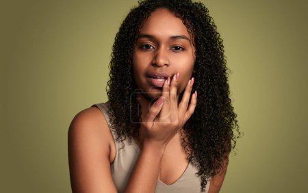 Foto de Hembra afro-americana positiva con cabello rizado tocando los labios y aplicando bálsamo labial curando herpes mientras está de pie sobre fondo amarillo en el estudio - Imagen libre de derechos