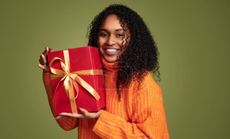 Photo for Cheerful african american girl in trendy orange sweater with curly hair, smiling and holding a big gift box over green background - Royalty Free Image