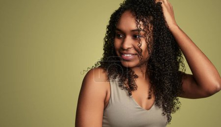 Téléchargez les photos : Femme afro-américaine positive en haut souriant et regardant loin tout en touchant les cheveux bouclés foncés sur fond vert - en image libre de droit