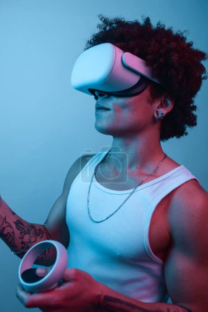 Photo for Casual African American male in goggles of virtual reality using controllers while playing game in neon light - Royalty Free Image