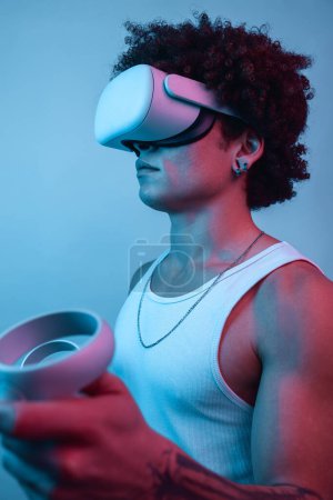 Photo for Vertical shot of Confident Hispanic male in VR goggles interacting with virtual reality against blue background in neon light - Royalty Free Image