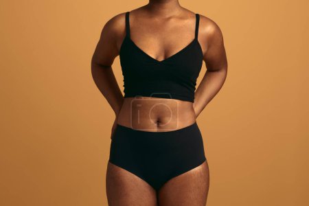 Téléchargez les photos : Modèle féminin afro-américain méconnaissable en lingerie noire debout sur fond beige avec les mains derrière le dos en studio - en image libre de droit