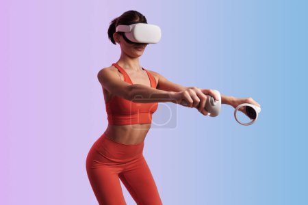 Téléchargez les photos : Adapter athlète féminine en vêtements de sport et casque VR faire squats tout en tenant les contrôleurs pendant l'entraînement de remise en forme sur fond de dégradé - en image libre de droit