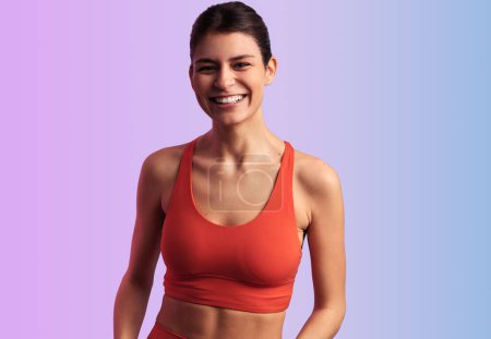 Photo for Portrait of happy slim Hispanic female athlete in orange crop top smiling and looking at camera, while standing against neon violet and blue background during fitness workout - Royalty Free Image