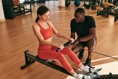 Photo for Full body high angle of young black instructor explaining technique of exercising on rowing machine, with strap to positive female in sportswear during fitness workout at gym - Royalty Free Image