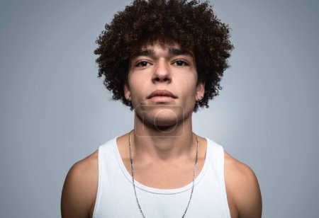 Téléchargez les photos : Headshot de gangster mâle brésilien dur hors la loi avec collier et boucles d'oreilles en tenue décontractée fixant la caméra sur fond gris - en image libre de droit