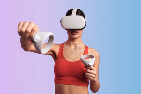 Photo for Confident young athletic woman in VR glasses and activewear practicing punches while playing videogame with joysticks against gradient background - Royalty Free Image