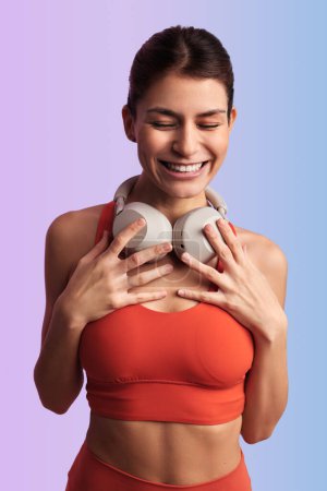 Photo for Happy Hispanic female athlete in sportswear with closed eyes holding earphones and smiling while standing against violet background and listening to music - Royalty Free Image