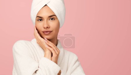 Photo for Young gentle female in white bathrobe and towel on head touching face tenderly after spa procedures looking at camera against pink background - Royalty Free Image