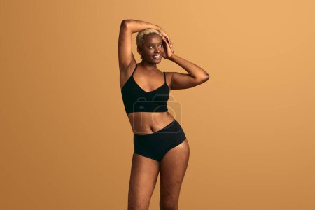 Foto de Mujer afroamericana sonriente curvilínea con pelo corto y rubio en lencería negra de pie sobre fondo beige con los brazos levantados y mirando hacia otro lado - Imagen libre de derechos