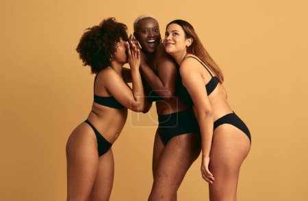 Téléchargez les photos : Joyeux jeunes modèles féminins multiraciaux en lingerie noire souriant et discutant de commérages ensemble tout en se tenant près les uns des autres sur fond beige en studio - en image libre de droit