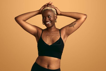 Téléchargez les photos : Jeune modèle féminin afro-américain souriant avec cheveux blonds afro touchant la tête portant des sous-vêtements noirs debout sur fond orange détournant les yeux - en image libre de droit