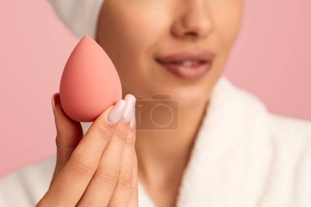Téléchargez les photos : Concentration douce de la récolte femelle anonyme en peignoir blanc démontrant mélangeur de maquillage pour appliquer le fond de teint sur fond rose - en image libre de droit