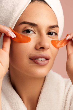 Photo for Young sensual female in bathrobe and towel on head applying moisturizing orange collagen patches under eyes during skincare routine against pink background - Royalty Free Image