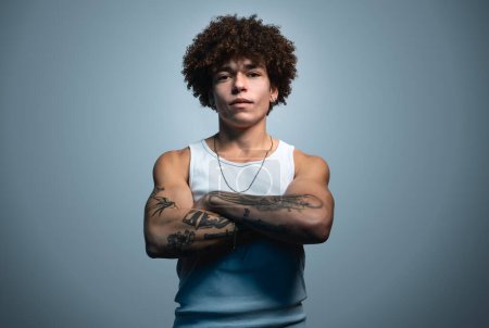 Foto de Portrait of vicious Brazilian male outlaw hooligan with curly brown hair and tattoos with necklace in casual wear gazing at camera against gray background - Imagen libre de derechos