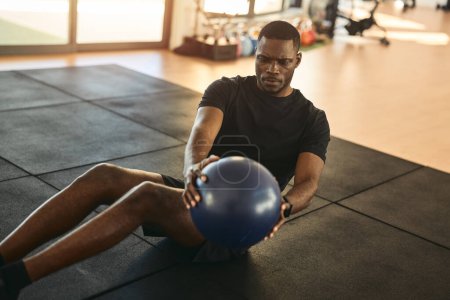 Téléchargez les photos : Jeune athlète masculin afro-américain fort en vêtements de sport noirs assis sur le tapis, faire de l'exercice de torsion russe avec une balle de médecine lourde pendant l'entraînement de remise en forme dans la salle de gym - en image libre de droit
