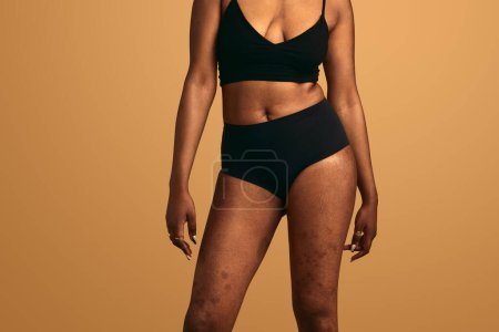 Téléchargez les photos : Crop anonyme modèle féminin africain en lingerie noire debout sur fond beige en studio - en image libre de droit