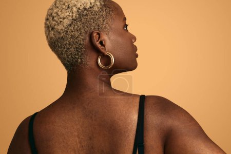 Foto de Vista posterior de la cosecha joven elegante modelo femenino afroamericano con el pelo corto teñido y la piel imperfecta mirando hacia otro lado contra el fondo beige - Imagen libre de derechos