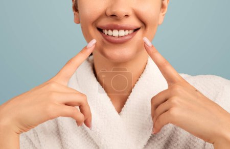 Photo for Crop ethnic lady with toothy smile in white bathrobe pointing at mouth on blue backdrop - Royalty Free Image