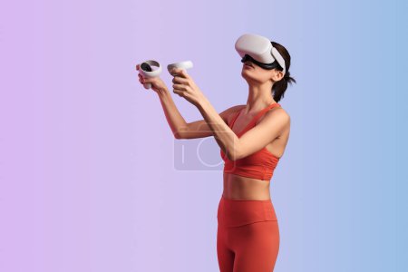 Photo for Young woman in red top leggings and VR headset with controllers playing videogame immersing in cyberspace against gradient background - Royalty Free Image