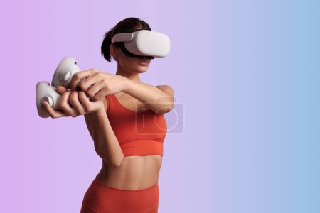 Téléchargez les photos : Adapter femelle dans les vêtements de sport et casque VR en utilisant des contrôleurs tout en jouant jeu vidéo interagissant avec la réalité invisible sur fond dégradé - en image libre de droit