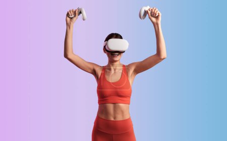 Photo for Happy young female gamer wearing red sportswear and VR headset raising arms with controllers while celebrating victory in videogame against gradient background - Royalty Free Image