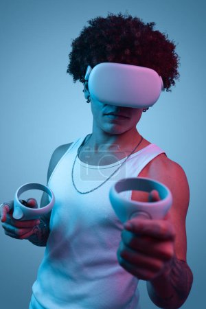 Photo for Concentrated young ethnic male millennial with curly Afro hair in white top and modern VR goggles, using controllers while playing video game against gray background - Royalty Free Image