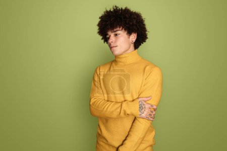 Foto de Confiado joven hispano con el pelo rizado oscuro y tatuaje en la mano en cuello alto amarillo abrazándose a sí mismo y mirando hacia otro lado contra el fondo verde - Imagen libre de derechos