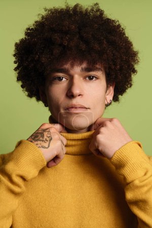 Foto de Joven modelo masculino étnico con cabello afro y tatuajes ajustando suéter de cuello alto amarillo mirando a la cámara mientras está de pie sobre fondo verde - Imagen libre de derechos