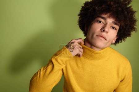 Foto de Retrato de un joven hispano con cabello afro y tatuajes en la mano tirando del cuello alto amarillo mientras mira a la cámara sobre fondo verde - Imagen libre de derechos
