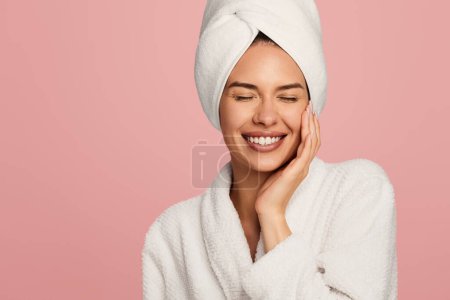 Foto de Mujer joven feliz en albornoz y toalla en la cabeza tocando la piel facial después del procedimiento de baño de pie sobre fondo rosa - Imagen libre de derechos