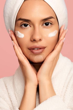 Téléchargez les photos : Cultiver les jeunes femmes portant un peignoir et une serviette sur la tête en appliquant de la crème hydratante sur les joues pendant le traitement de la peau en studio sur fond rose - en image libre de droit