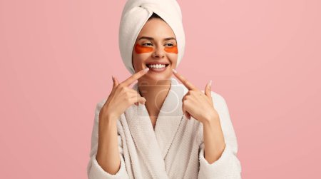 Foto de Mujer joven optimista con parches para los ojos que usan albornoz y toalla en la cabeza que señala la sonrisa de los dientes contra el fondo rosa - Imagen libre de derechos