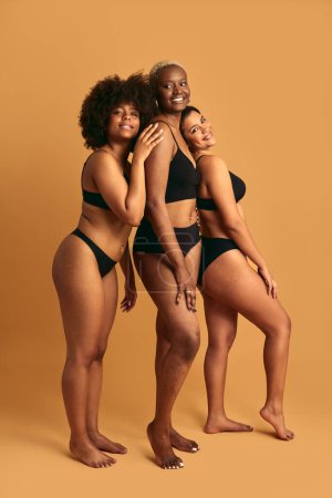 Téléchargez les photos : Vue latérale pleine longueur de modèles féminins multiraciaux aux pieds nus positifs en sous-vêtements noirs, souriants et regardant la caméra tout en se tenant debout sur un fond beige - en image libre de droit