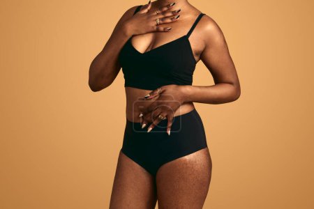 Photo for Crop unrecognizable African American female model with stretch marks on hips in black underwear touching body while standing in studio against beige background - Royalty Free Image