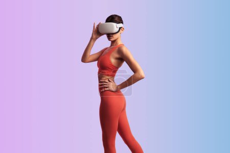 Foto de Vista lateral de mujer delgada en gafas VR y ropa deportiva de pie con la mano en la cintura y explorando el ciberespacio contra el fondo degradado - Imagen libre de derechos