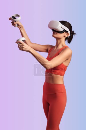 Photo for Young slim sportswoman in red activewear and VR headset raising hands with controllers while playing videogame exploring virtual world against gradient background - Royalty Free Image