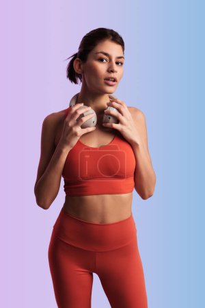 Téléchargez les photos : Jeune athlète féminine confiante en vêtements de sport avec écouteurs sans fil regardant la caméra tout en se tenant debout sur fond violet - en image libre de droit