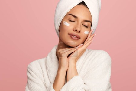 Photo for Hispanic female in bathrobe and towel on head closing eyes while having cream smeared on cheeks against pink background - Royalty Free Image