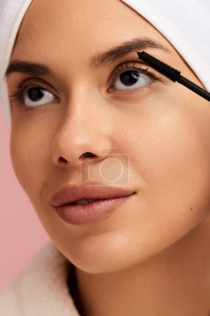 Photo for Crop young female model with perfect smooth skin wearing white bathrobe and towel on head, applying black mascara with brush on eyelashes during beauty routine - Royalty Free Image