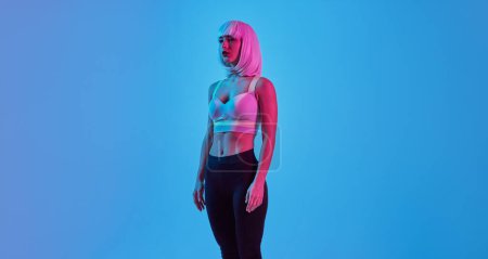 Foto de Futurista joven cyborg femenino en traje de moda y peluca mirando hacia otro lado mientras está de pie sobre fondo azul en luz de neón - Imagen libre de derechos