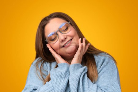 Photo for Positive young overweight female in glasses smiling and touching cheeks against yellow background - Royalty Free Image